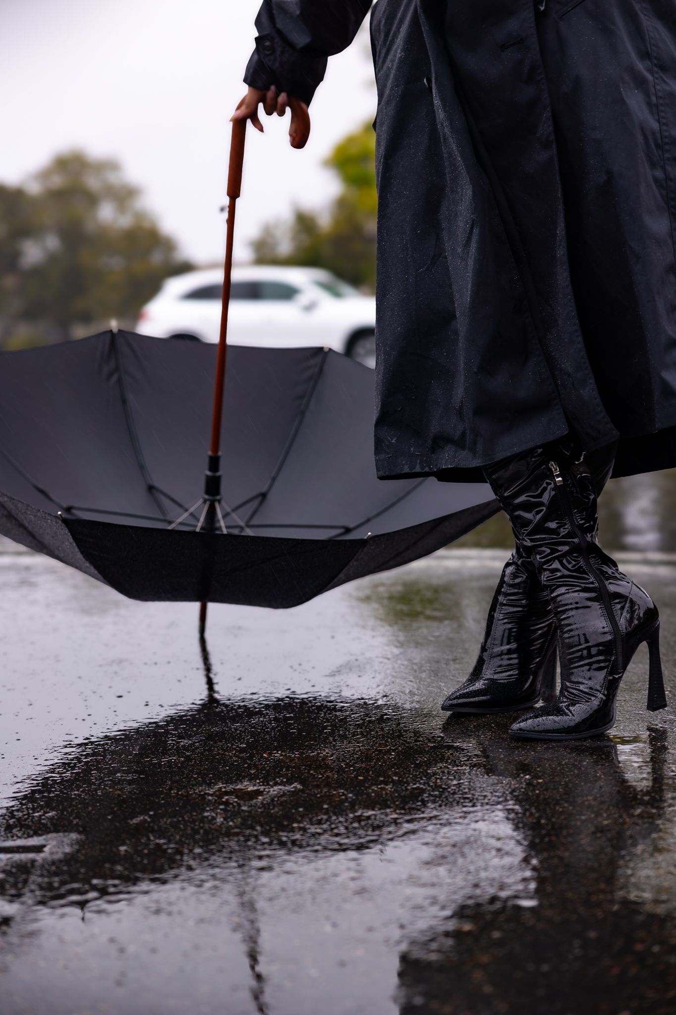 Close-up of black boots and umbrella reflecting in wet pavement during rainfall.