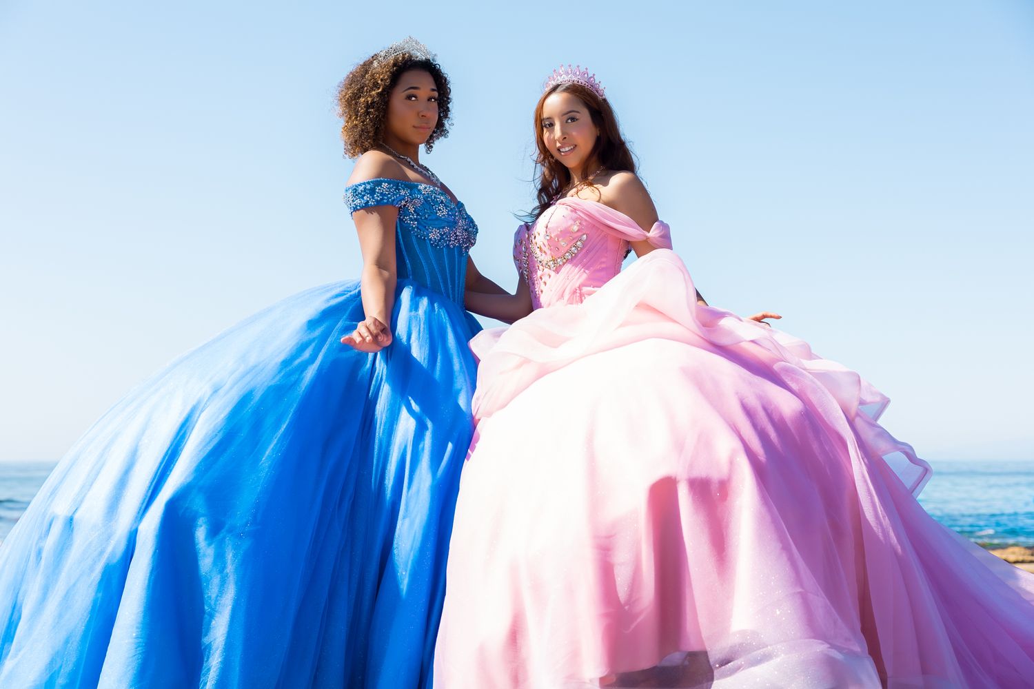 Two friends in elegant ballgowns, one in royal blue and one in pink, pose together against a bright sky.