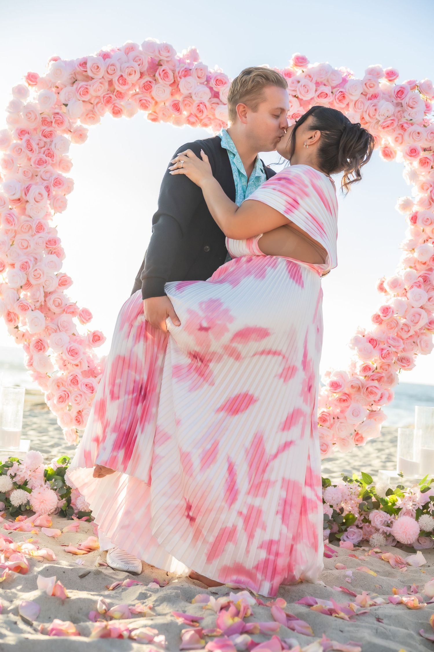 Romantic beach proposal scene with pink floral decorations and city skyline in background.