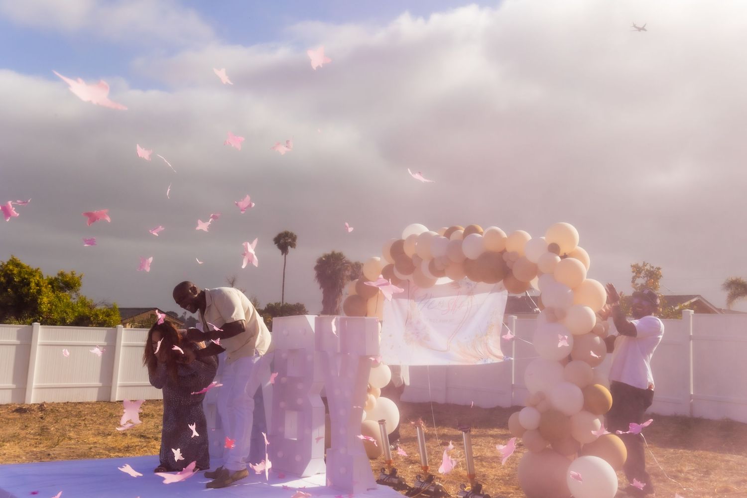 Outdoor celebration with pink flower petals and balloons being released into cloudy sky.