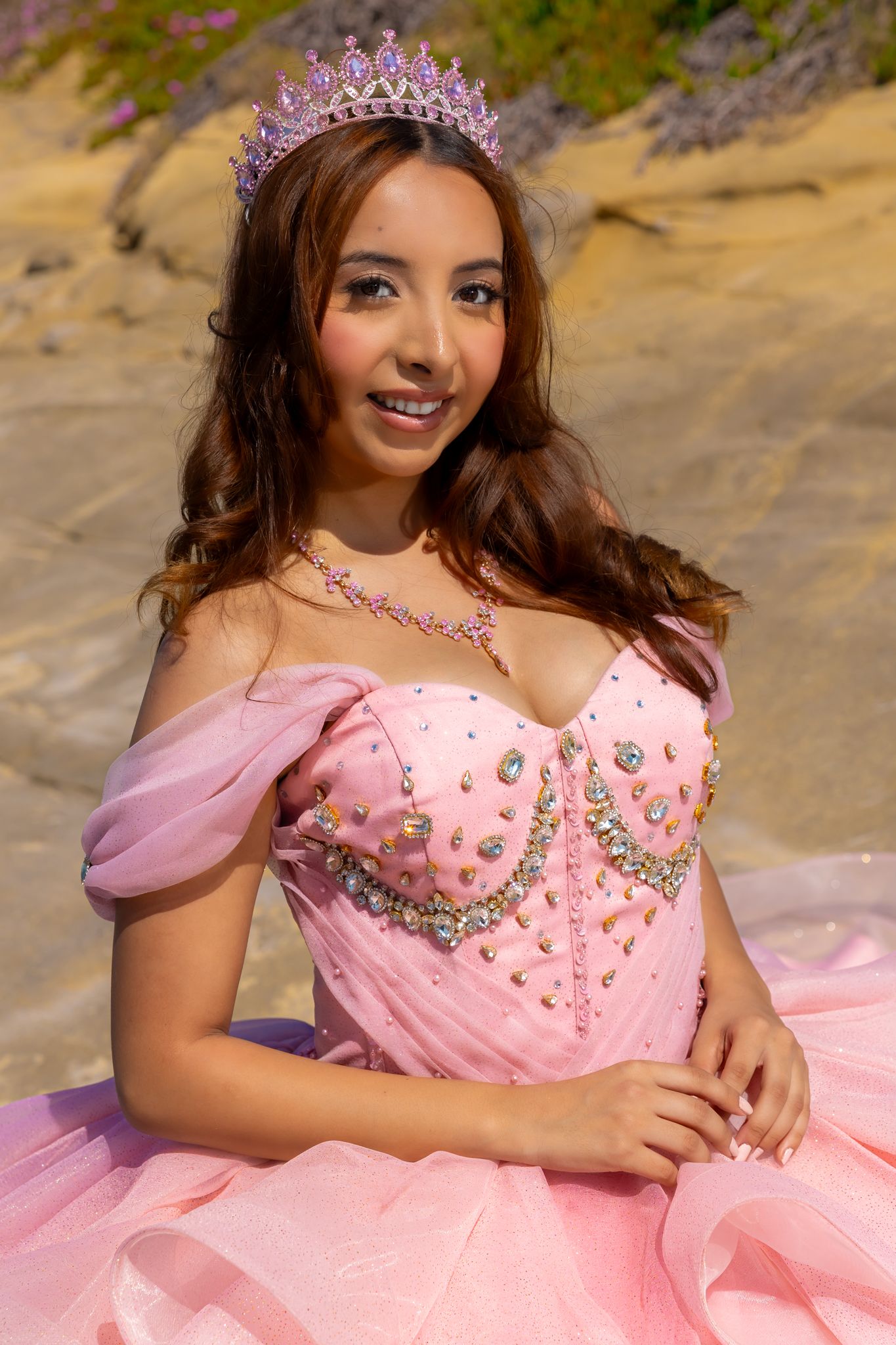 A person in a pink jeweled gown and tiara poses outdoors with natural scenery in the background.