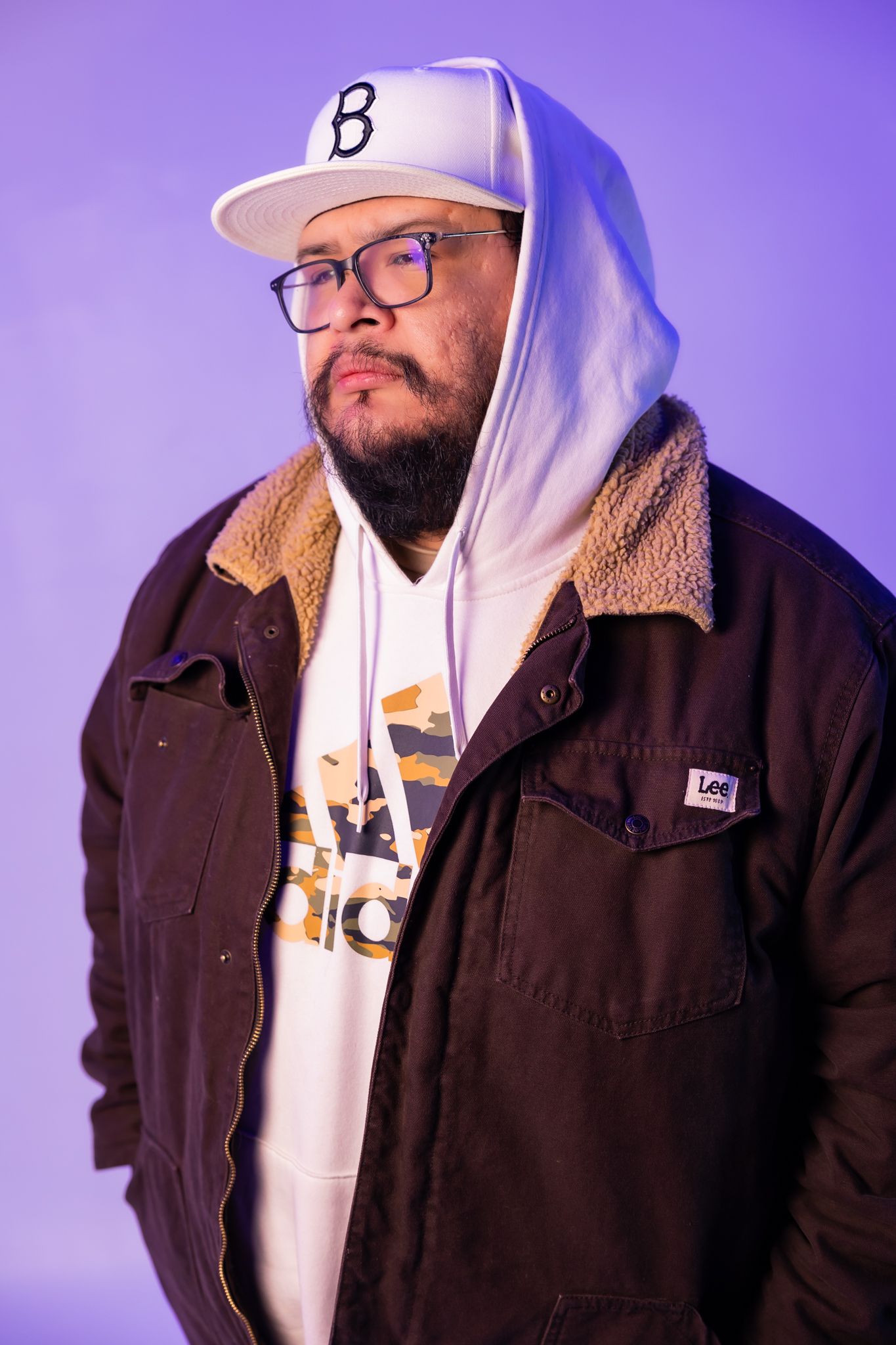 Portrait with Brooklyn cap, glasses and brown jacket against purple background.