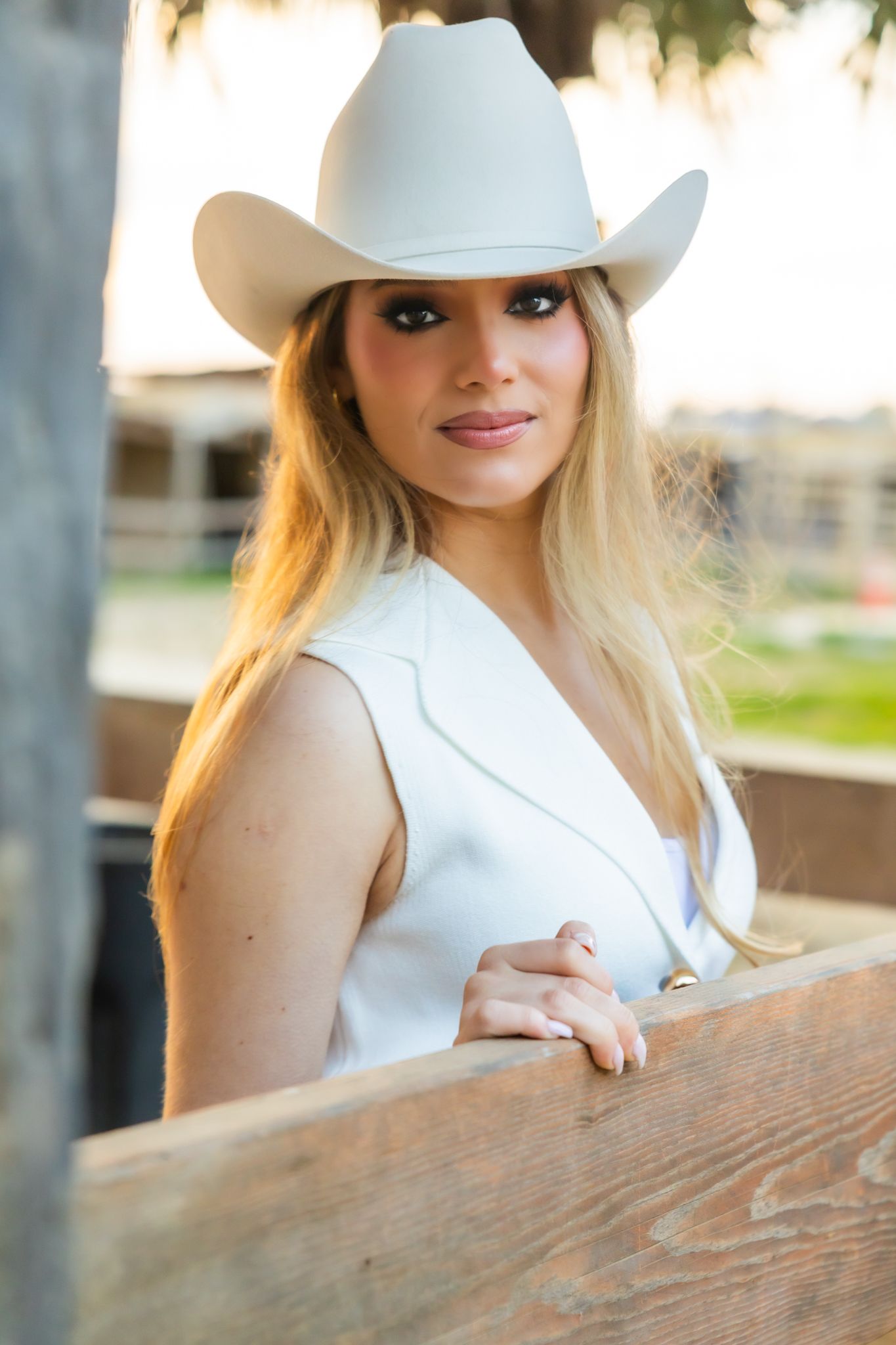 Person wearing a sleeveless white top and cowboy hat posing against a rustic wooden fence.