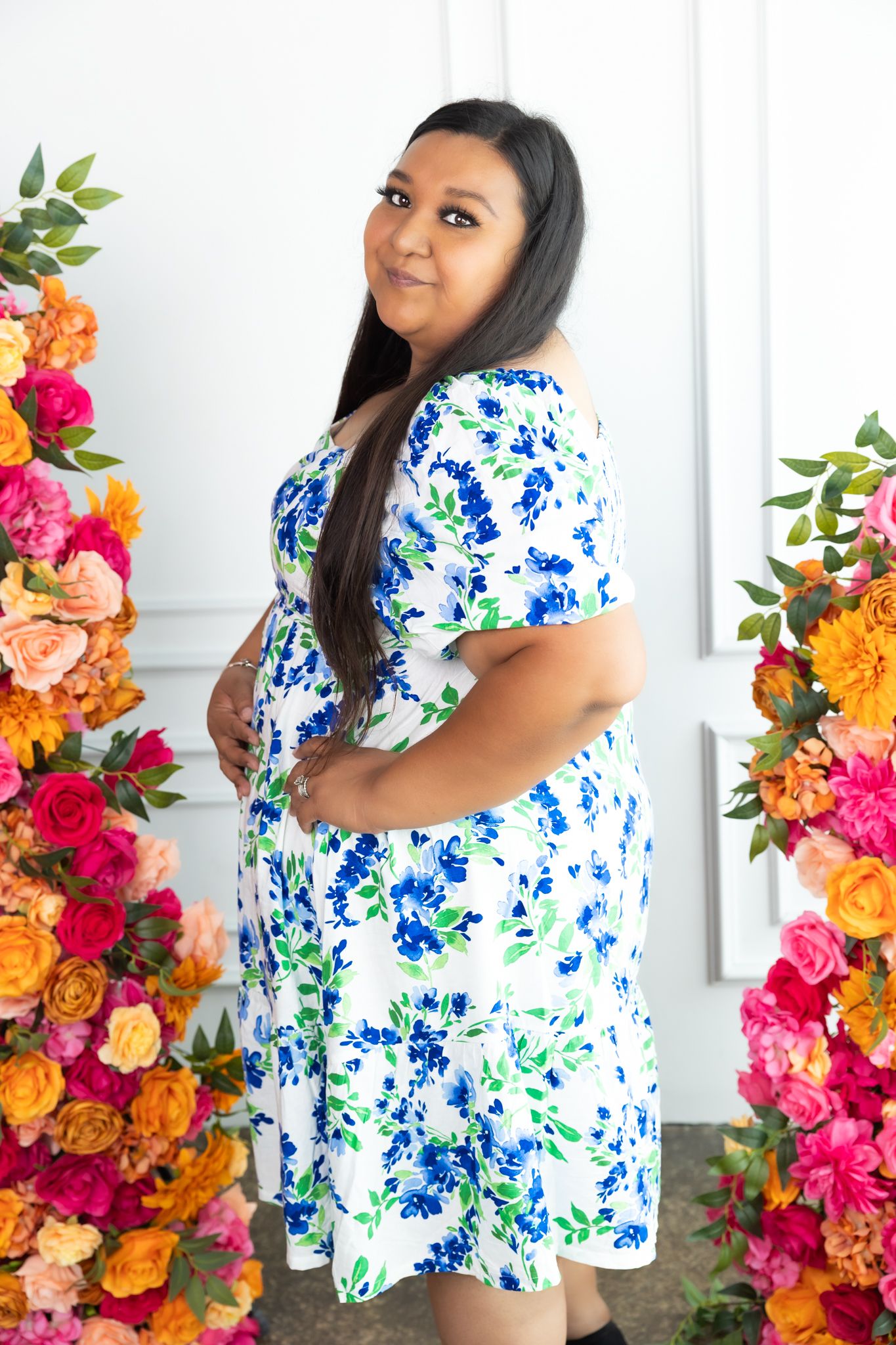 Woman in a blue and white floral dress poses between colorful flower arrangements.
