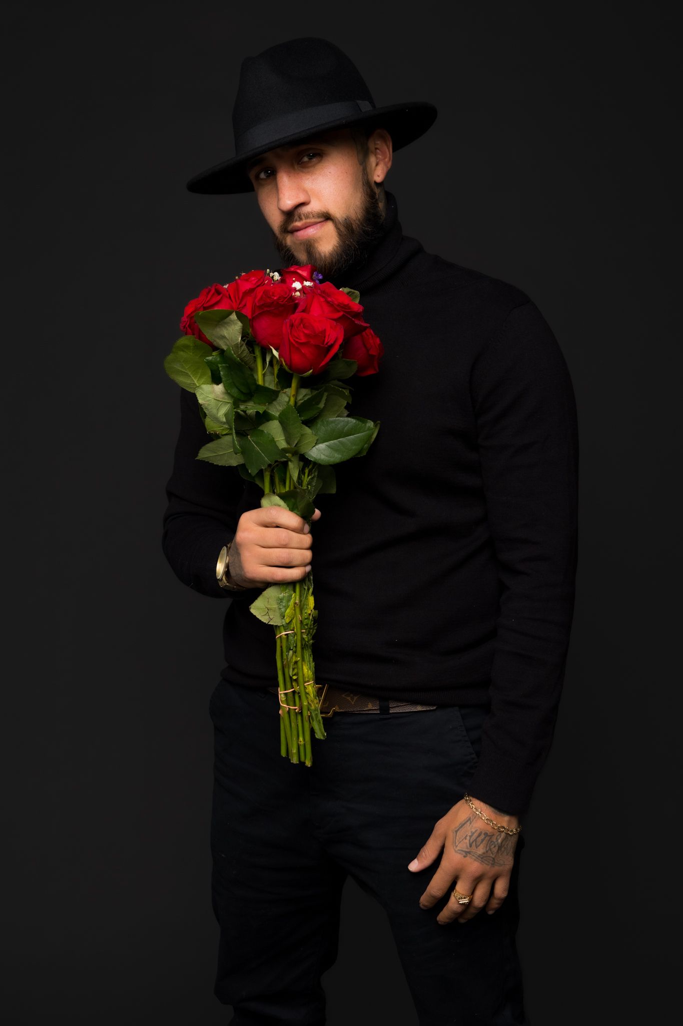Figure in black holding red roses against dark background in dramatic lighting.