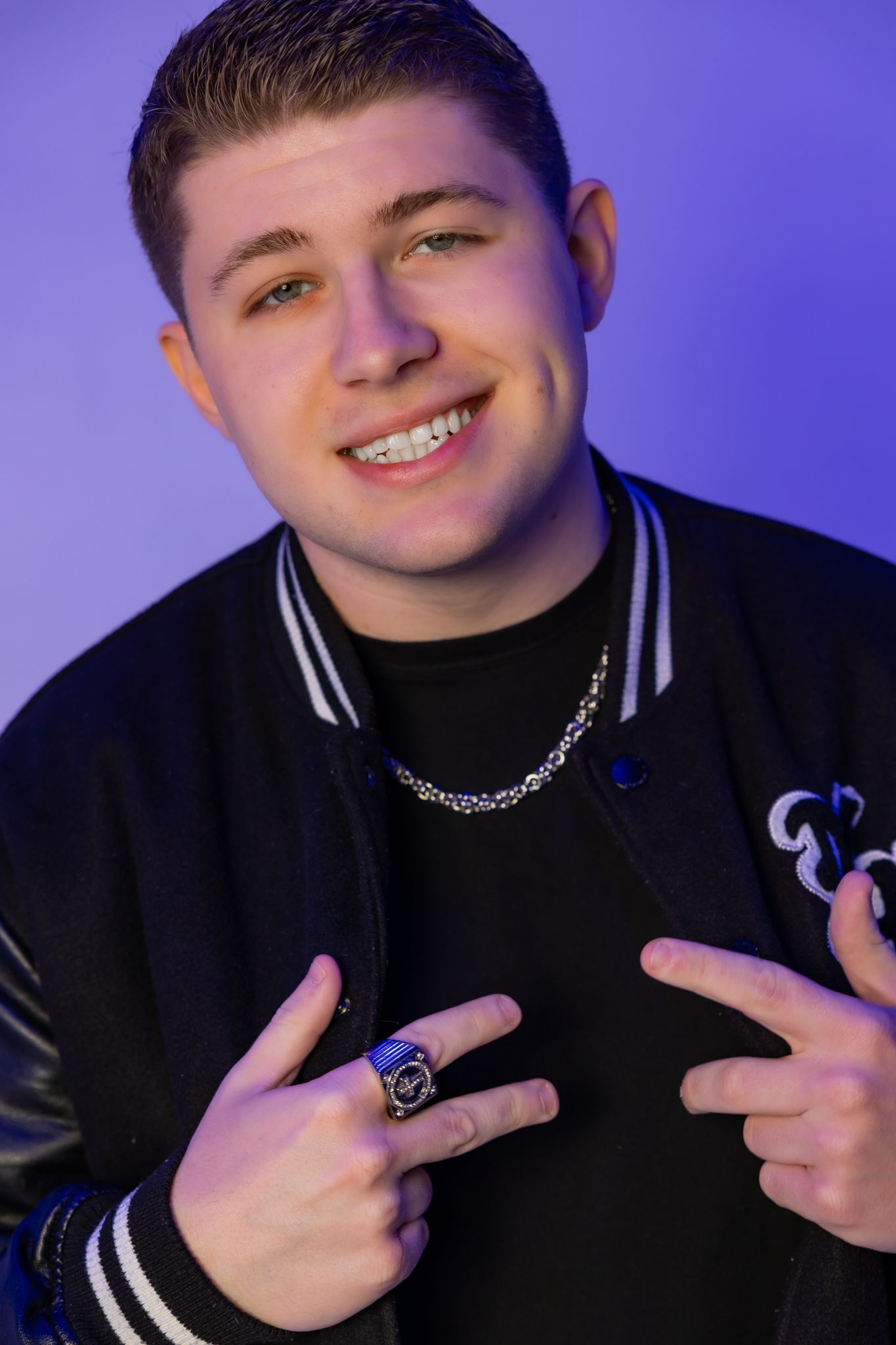 Person in black varsity jacket poses with hand gestures against purple studio background.
