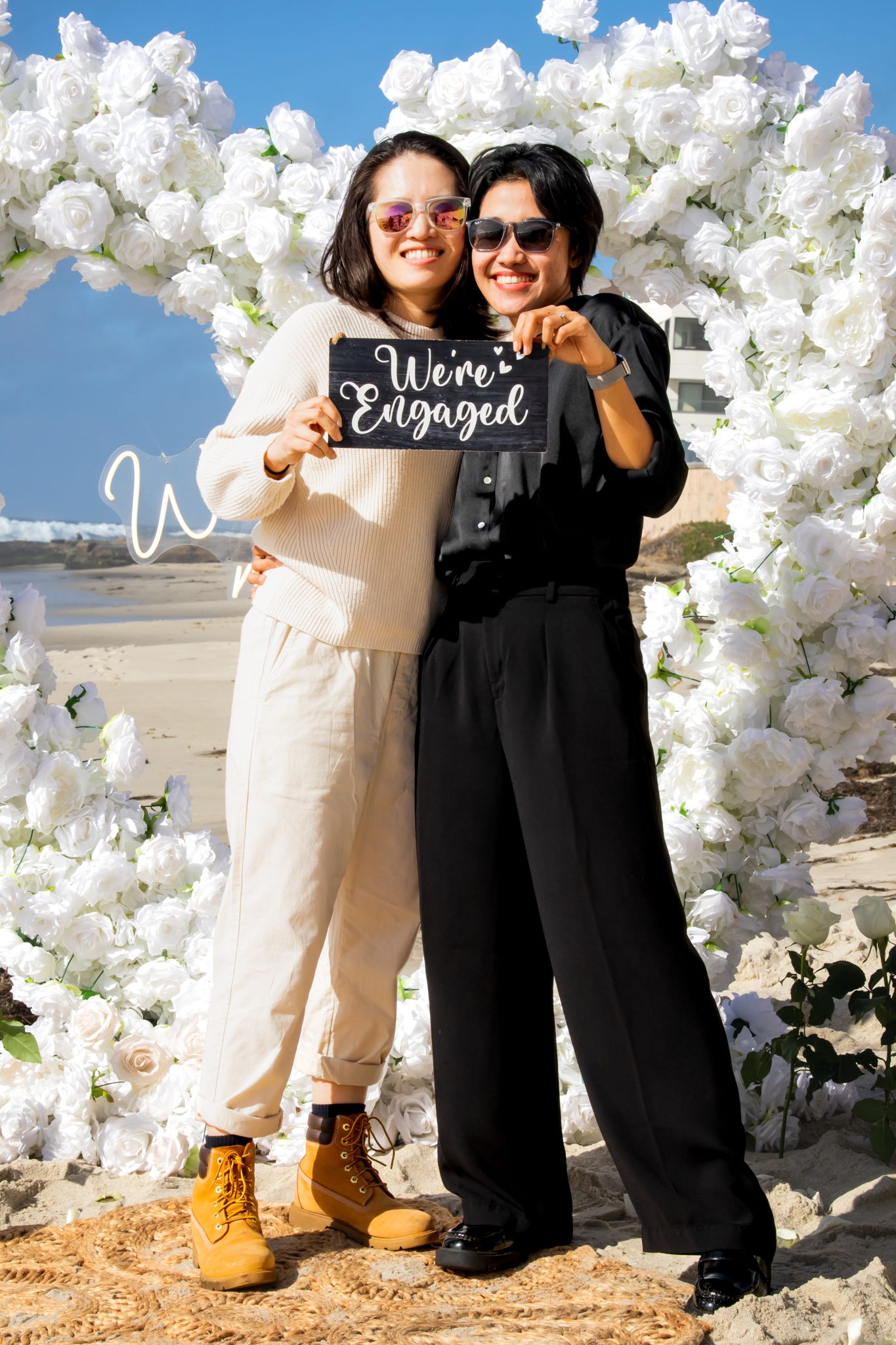 Two people pose together under an elegant white balloon and flower arch by the oceanside.