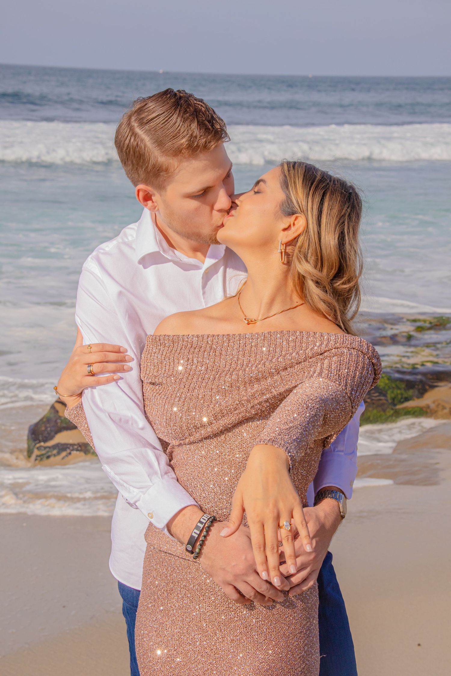 Couple in formal attire embracing on a scenic beach during sunset with waves in the background.