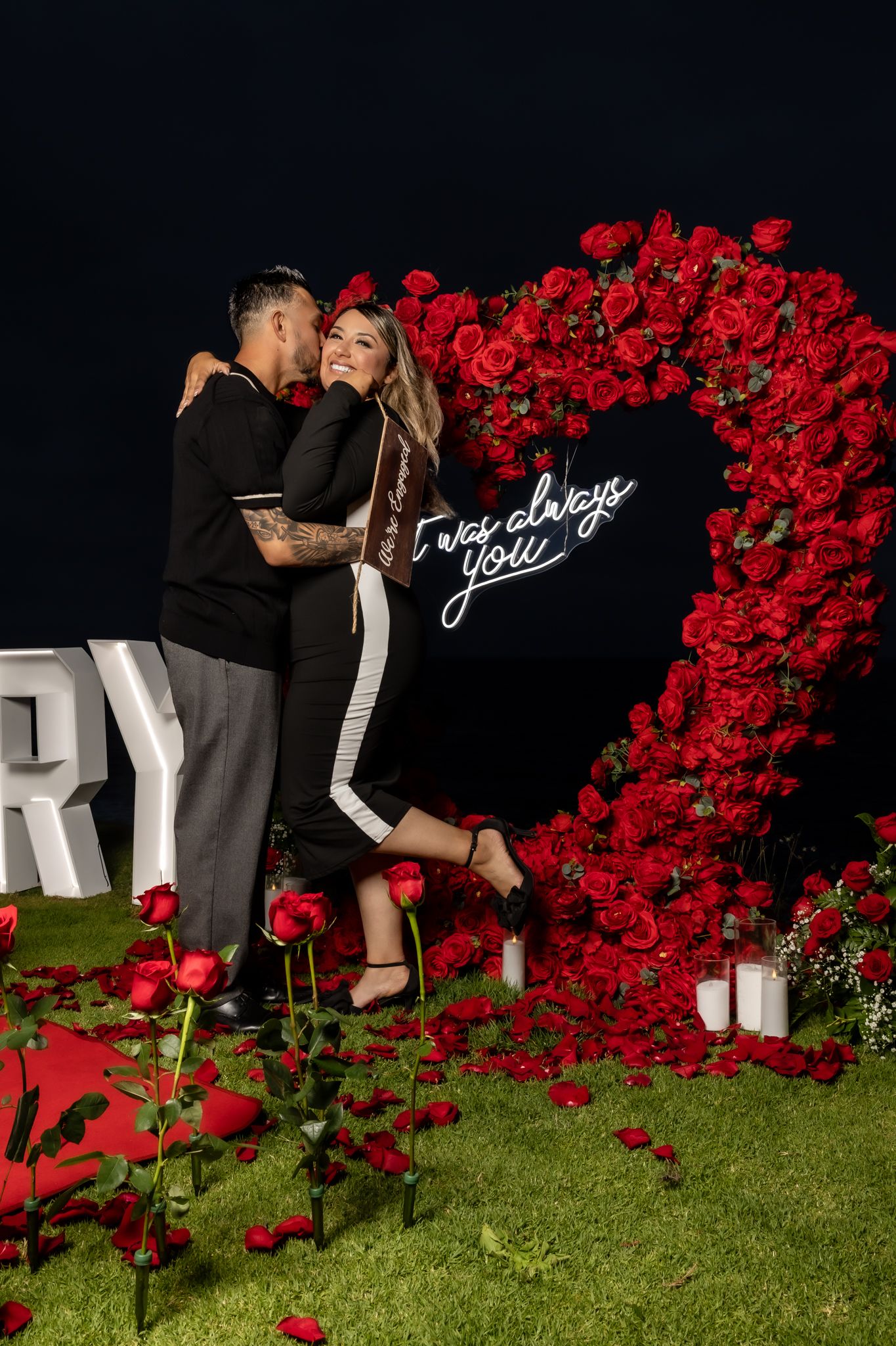 Couple posing against black wall decorated with red roses and neon signage.