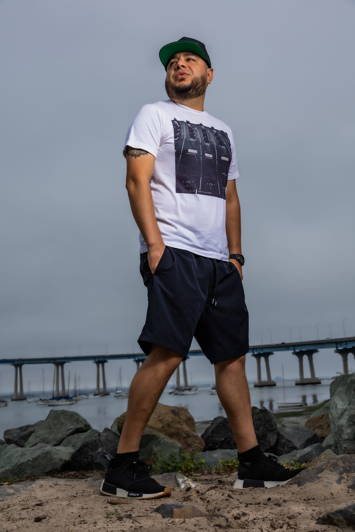 Person wearing white t-shirt and black shorts posing on rocky beach with pier in background.