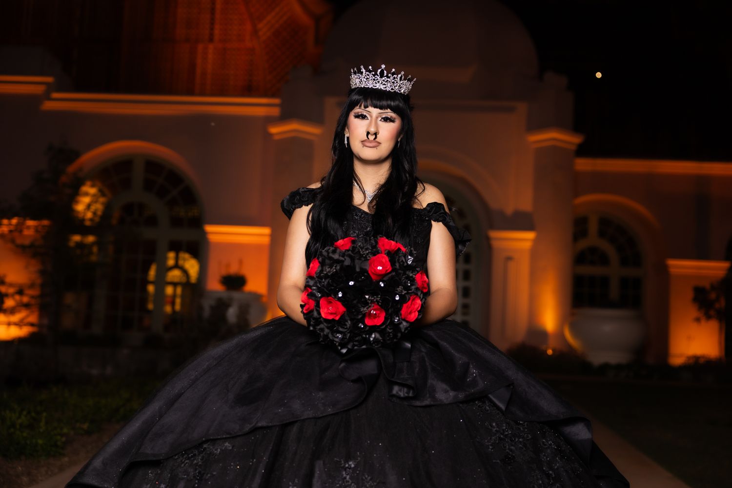 A gothic-styled quinceañera dress in black with red roses photographed at night with illuminated architecture.