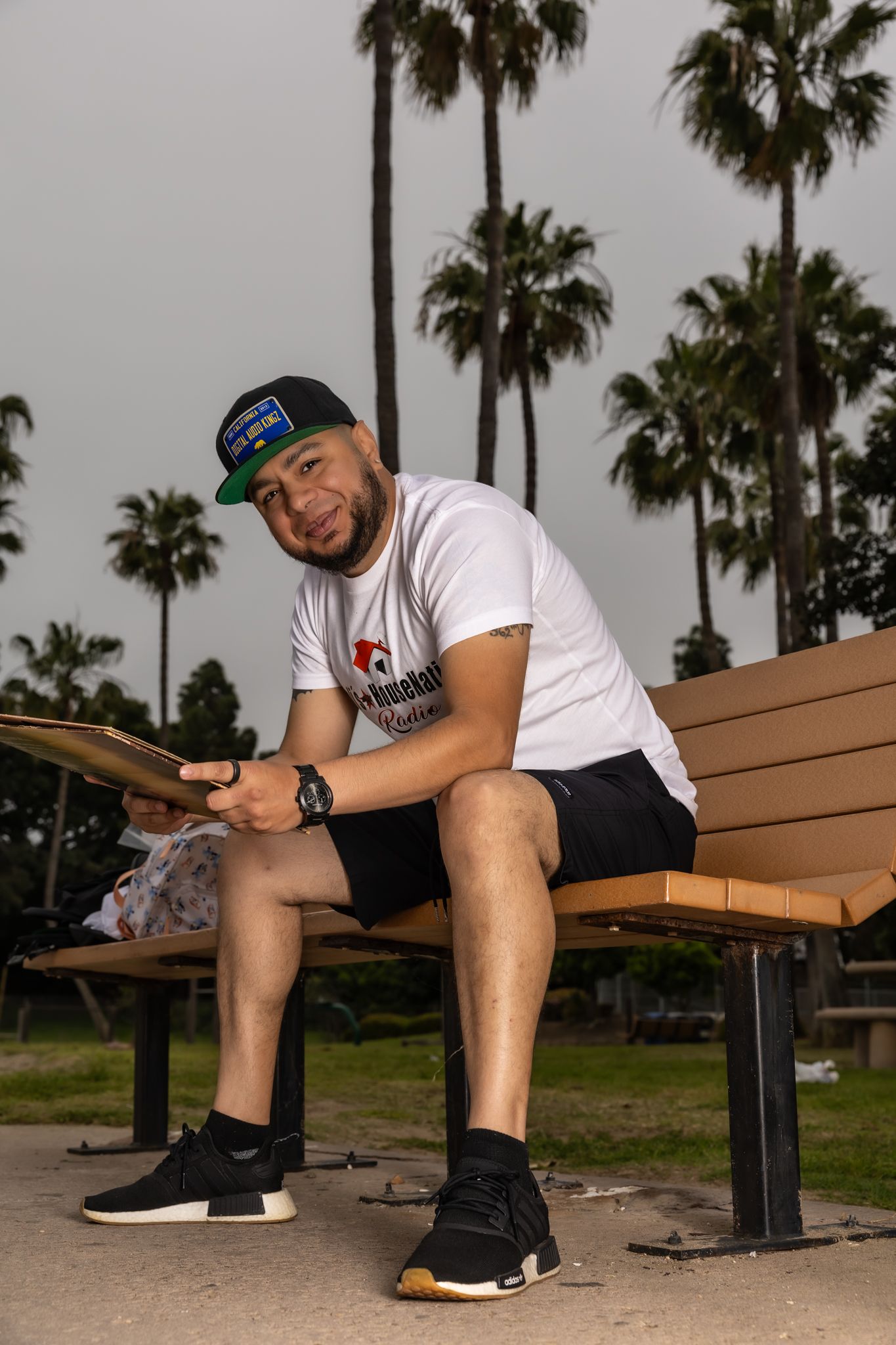 Person sitting on wooden bench wearing casual attire and baseball cap near palm trees.
