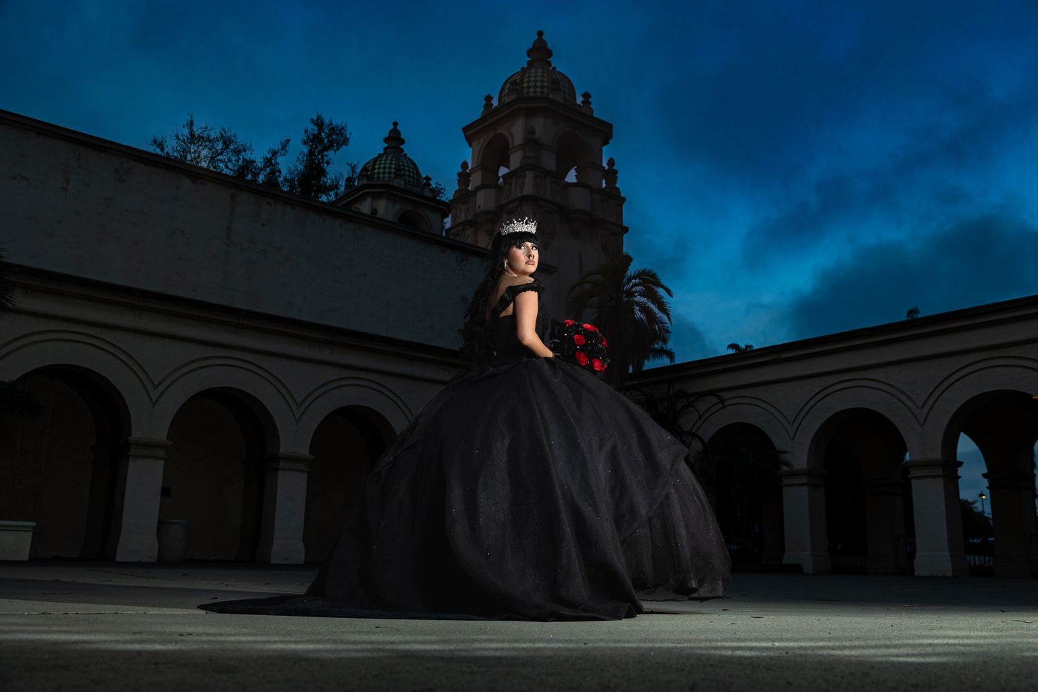 A dramatic black ballgown silhouette against historic architecture and dark blue evening sky.