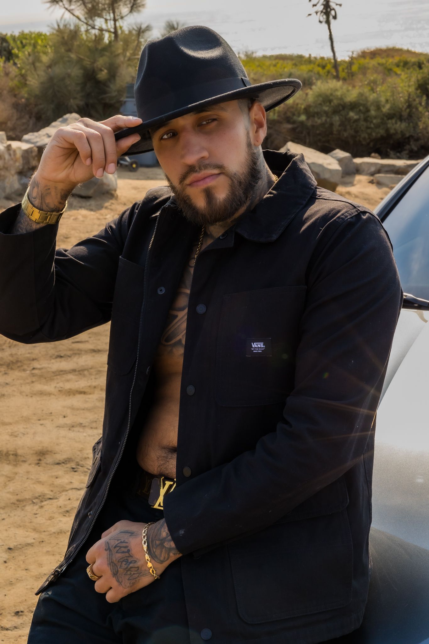 A stylish portrait in a desert setting featuring a dark jacket, fedora hat, and visible tattoos against a natural backdrop.