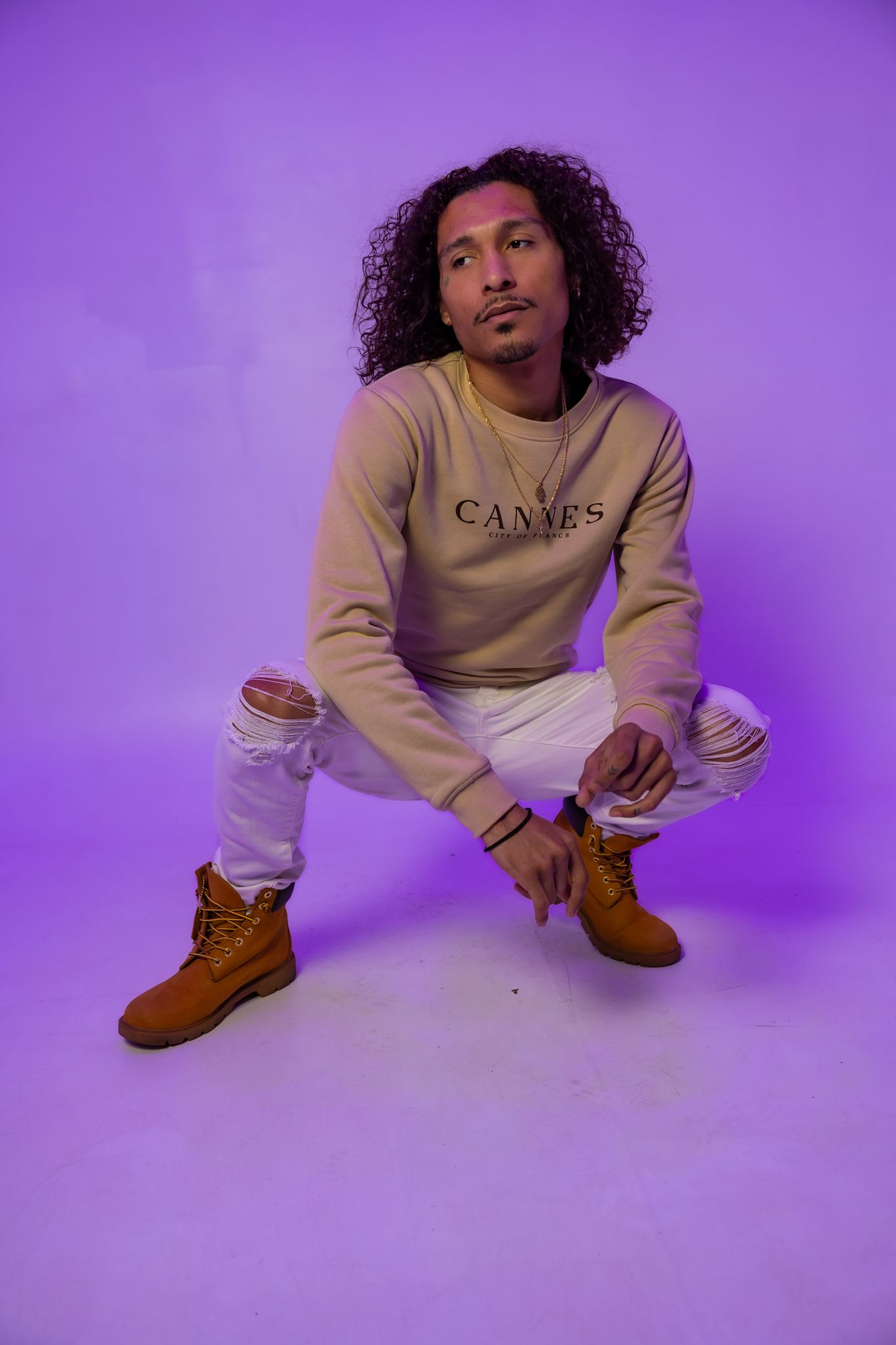 Person in beige CANNES sweatshirt and brown boots poses on white floor with purple lighting.