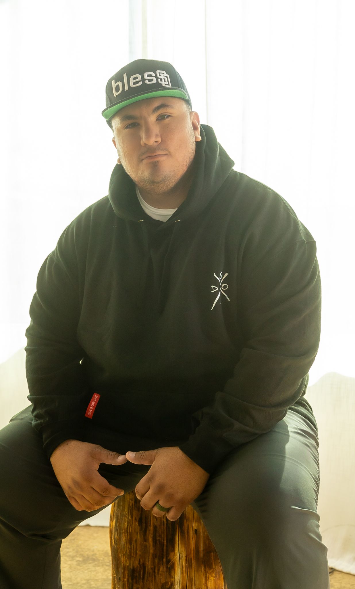 Individual in a green cap and black hoodie sitting casually on a wooden surface in natural lighting.