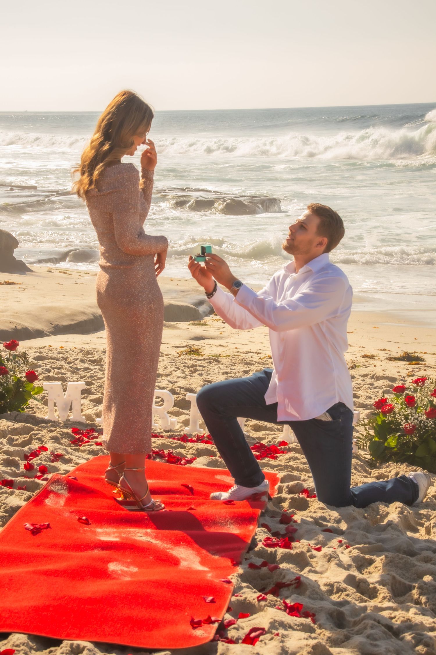 A romantic beach proposal setup with red rose petals on a blanket as a couple gets engaged by the ocean waves.