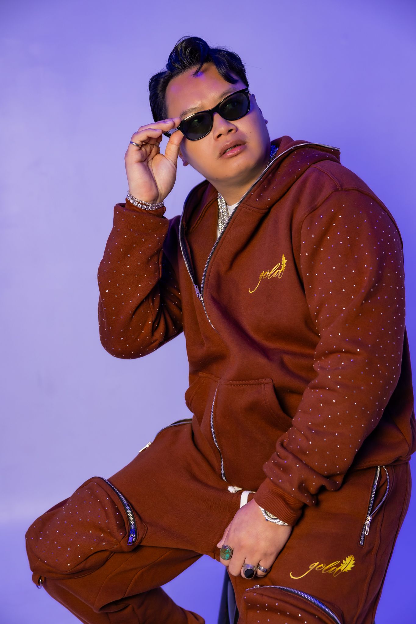 Model wearing brown sparkly tracksuit poses confidently against purple background.