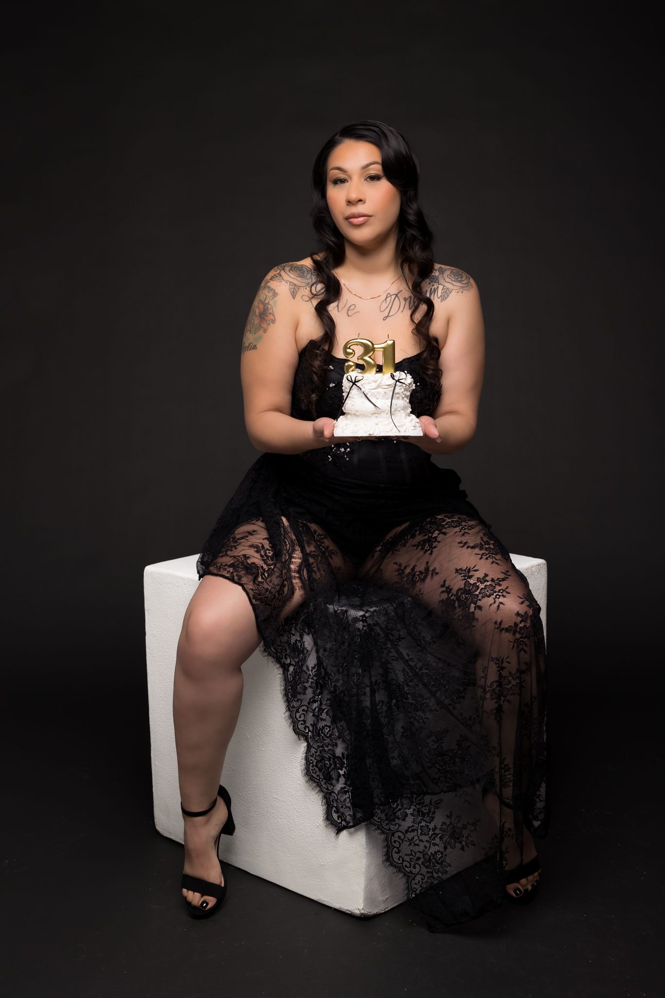 Elegant boudoir portrait featuring a black lace dress and white cube seat against dark background.