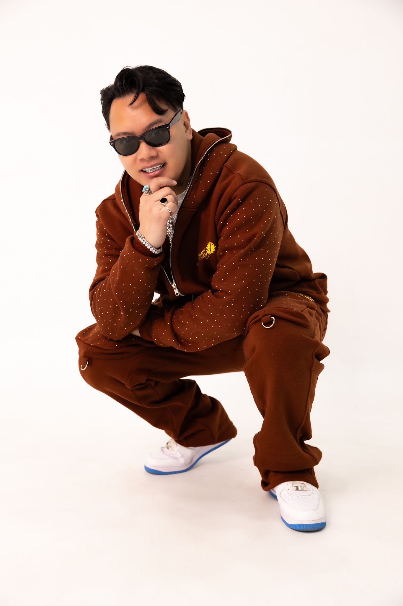 Portrait of person in brown teddy fleece tracksuit and sunglasses crouching against white background.