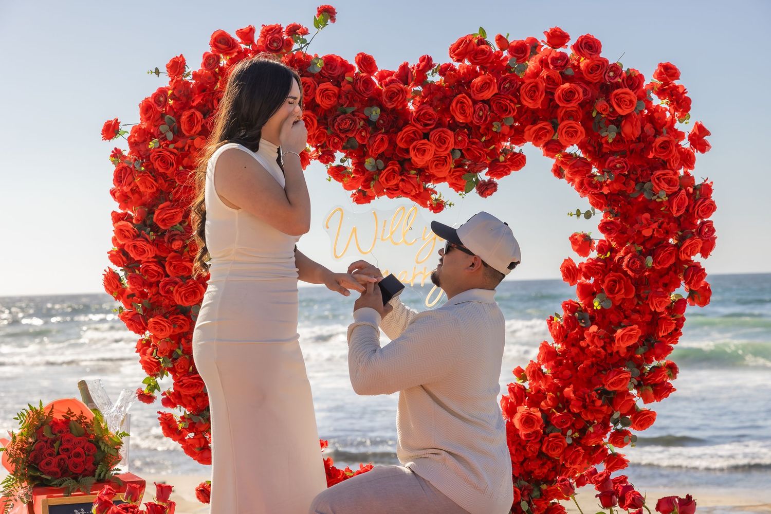 Beautiful beach proposal setting with a heart-shaped arch made of red roses against ocean backdrop.