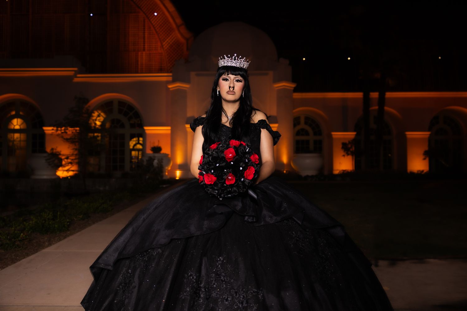 A black ballgown with red floral accents photographed at night with warm architectural lighting.