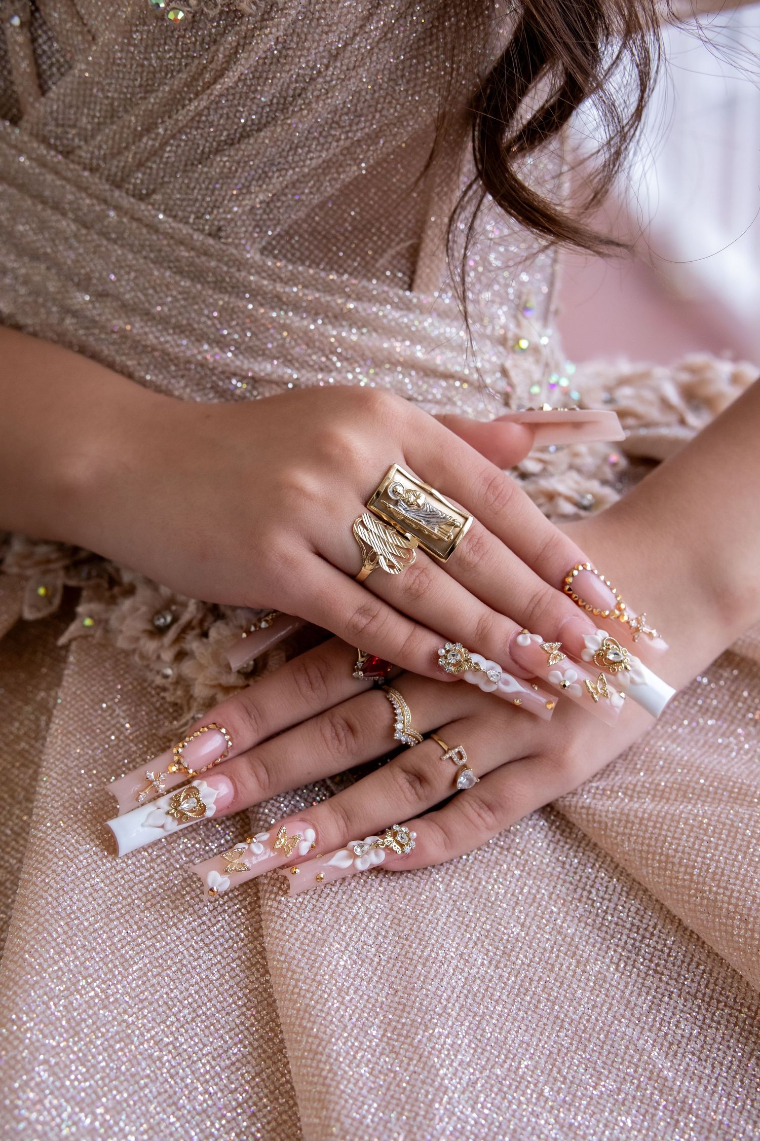Elaborately decorated long acrylic nails and rings displayed on a sparkly champagne-colored dress fabric.