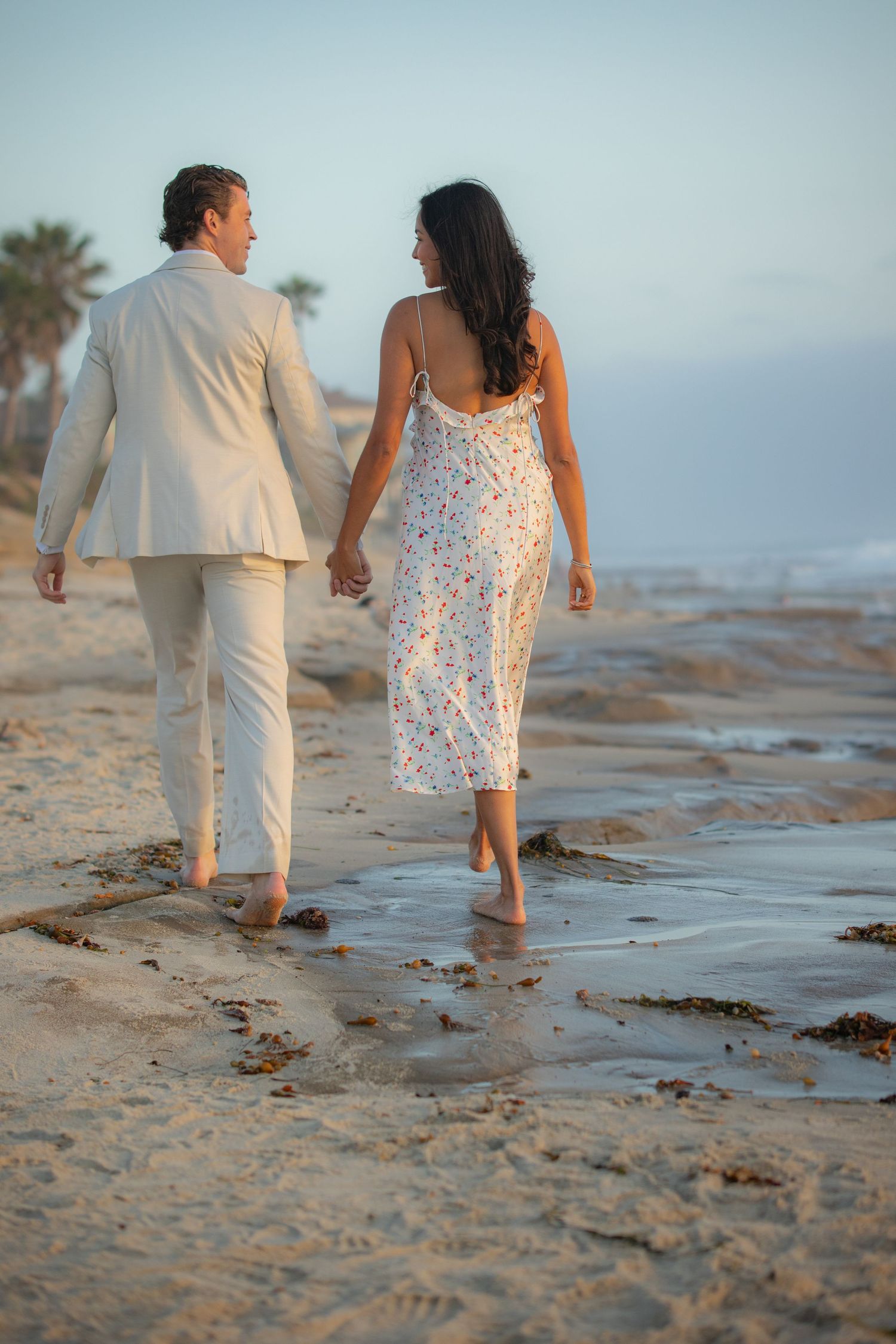 A couple walks hand in hand along a beach shoreline during sunset wearing white and floral outfits.