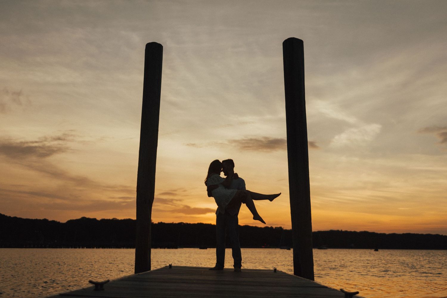 Silhouetted couple embracing on dock at sunset with dramatic sky and water backdrop.
