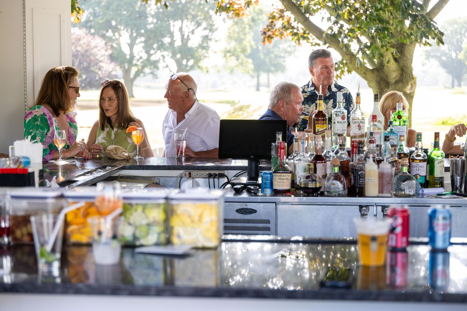 Guests enjoy drinks at an outdoor bar stocked with bottles at a sunny event venue.