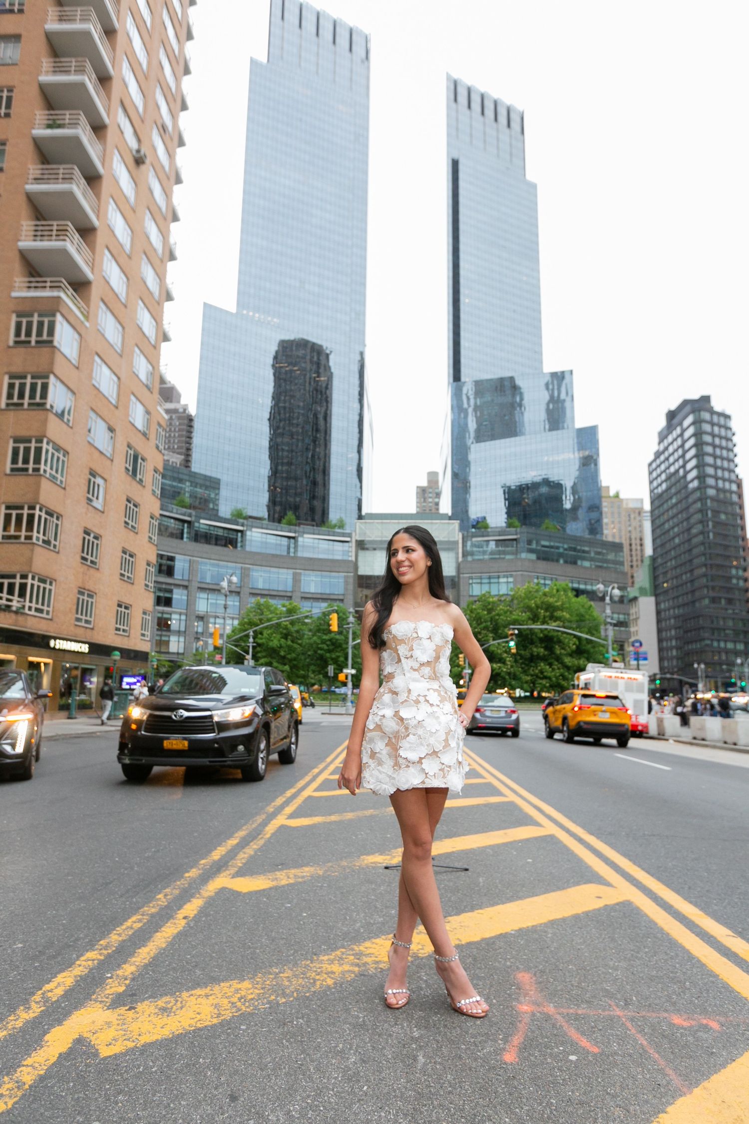 Woman in white floral mini dress and heels posing on NYC street with skyscrapers in background.