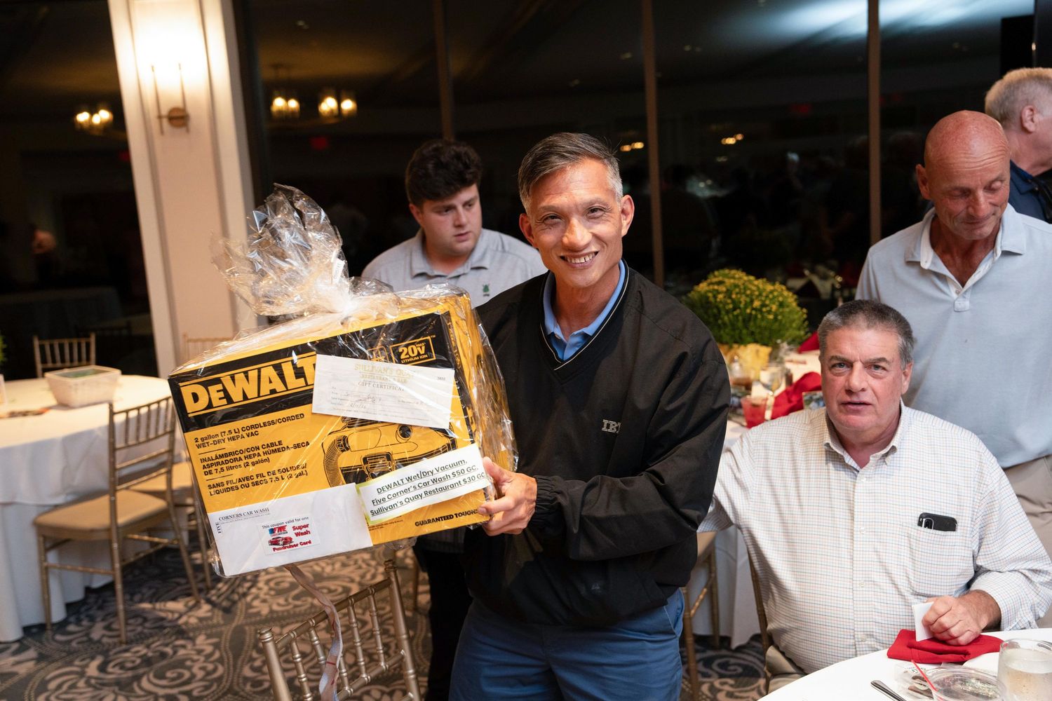 Smiling man holds a DeWalt tool set prize at an indoor charity golf tournament dinner event.