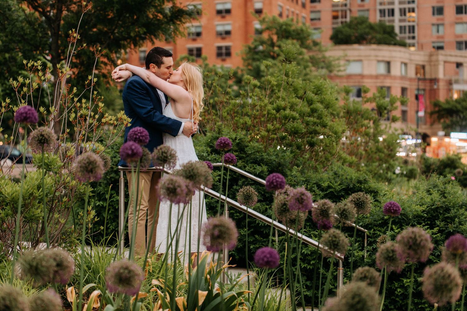 Couple embracing among tall purple allium flowers in urban garden setting.