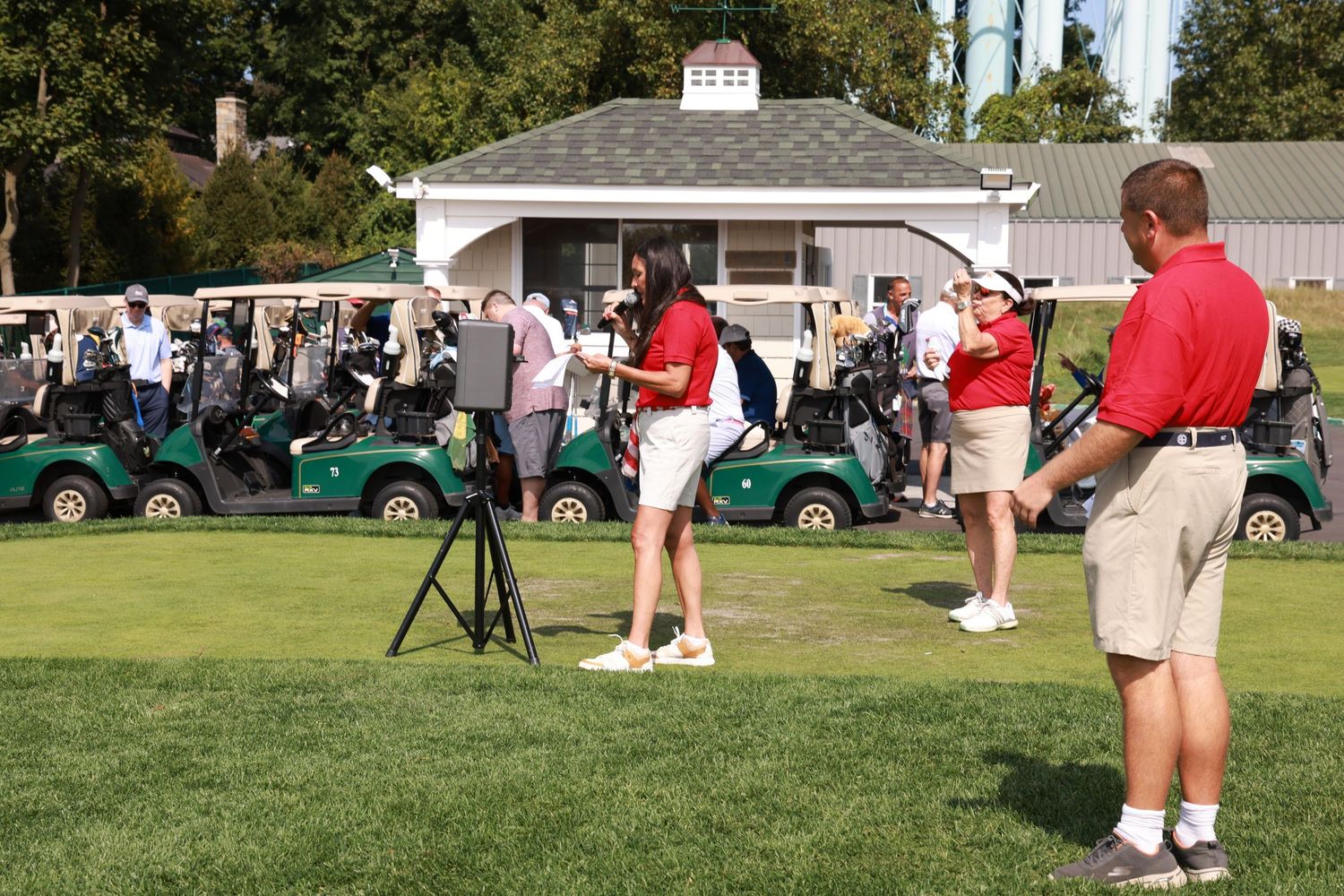 Golfers and staff gather on a green fairway near golf carts at an outdoor golf tournament event.
