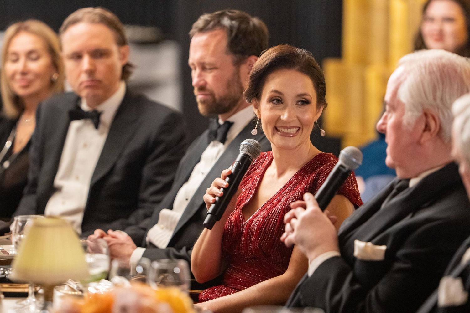 Woman in red dress speaks at formal gala dinner event with guests in evening wear seated at decorated table.
