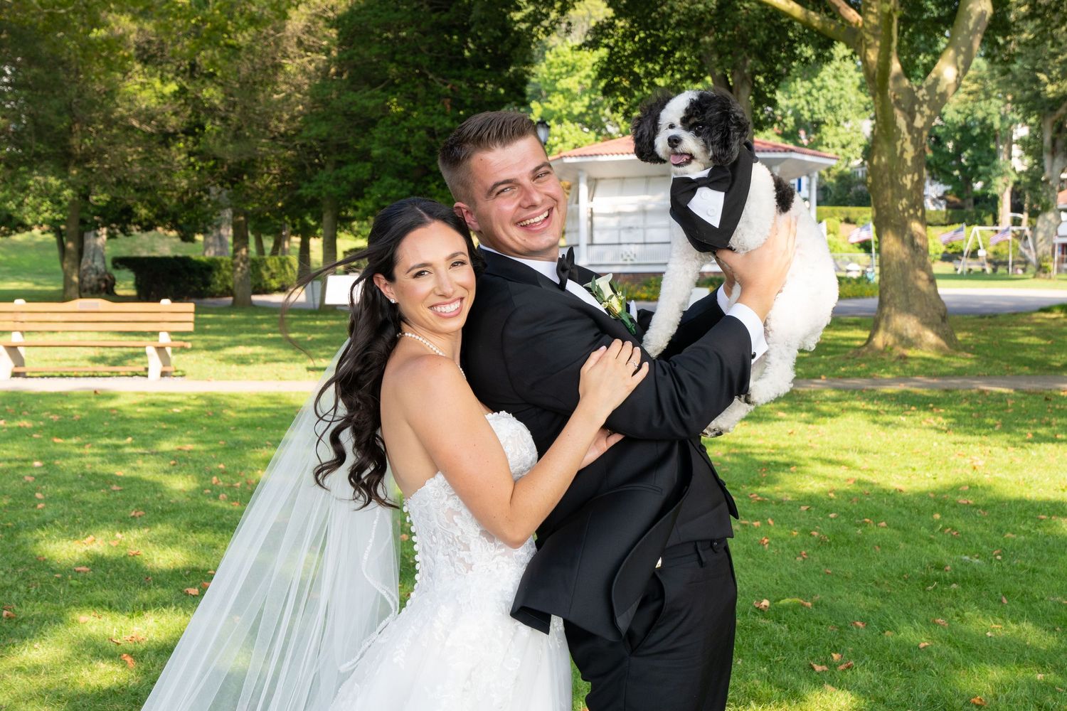 A couple poses joyfully with their dog in a sunny garden setting.