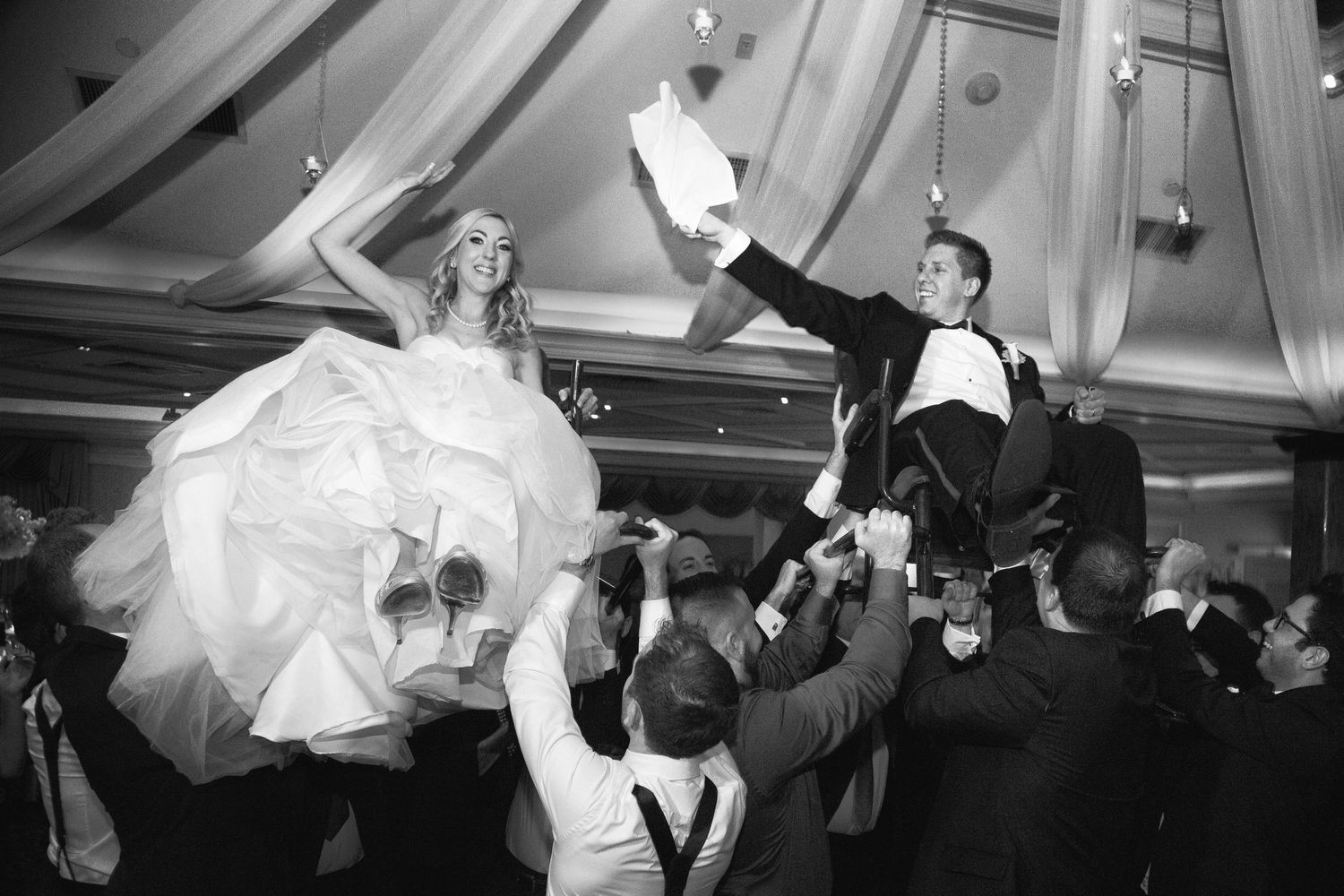 Wedding guests lift the bride and groom on chairs during a lively reception celebration.