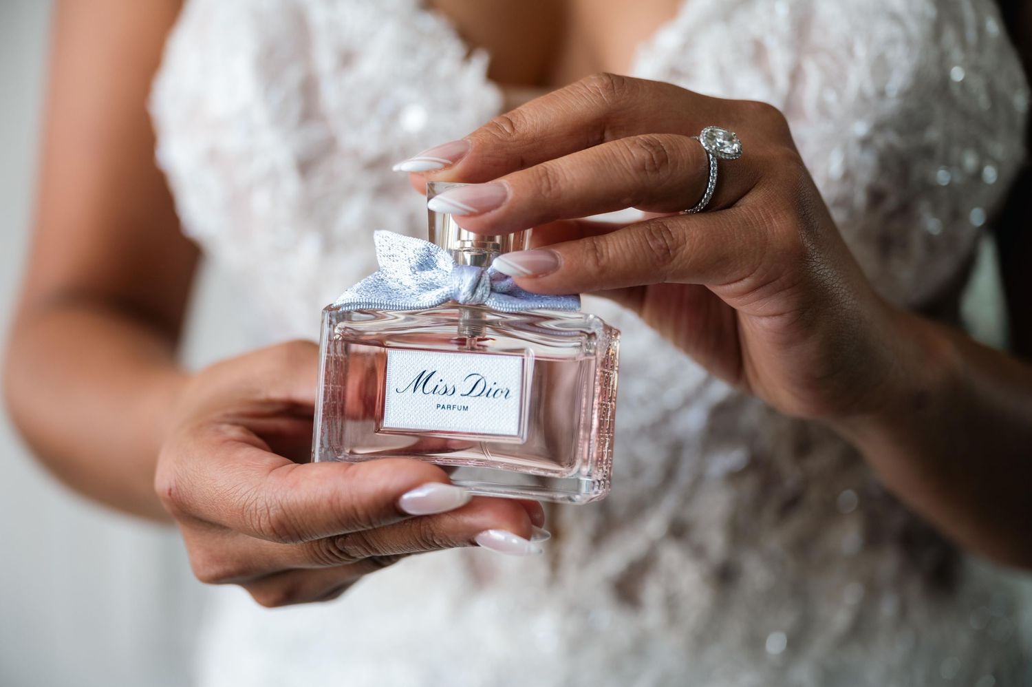 Close-up of hands holding a Miss Dior perfume bottle in delicate lace wedding attire.