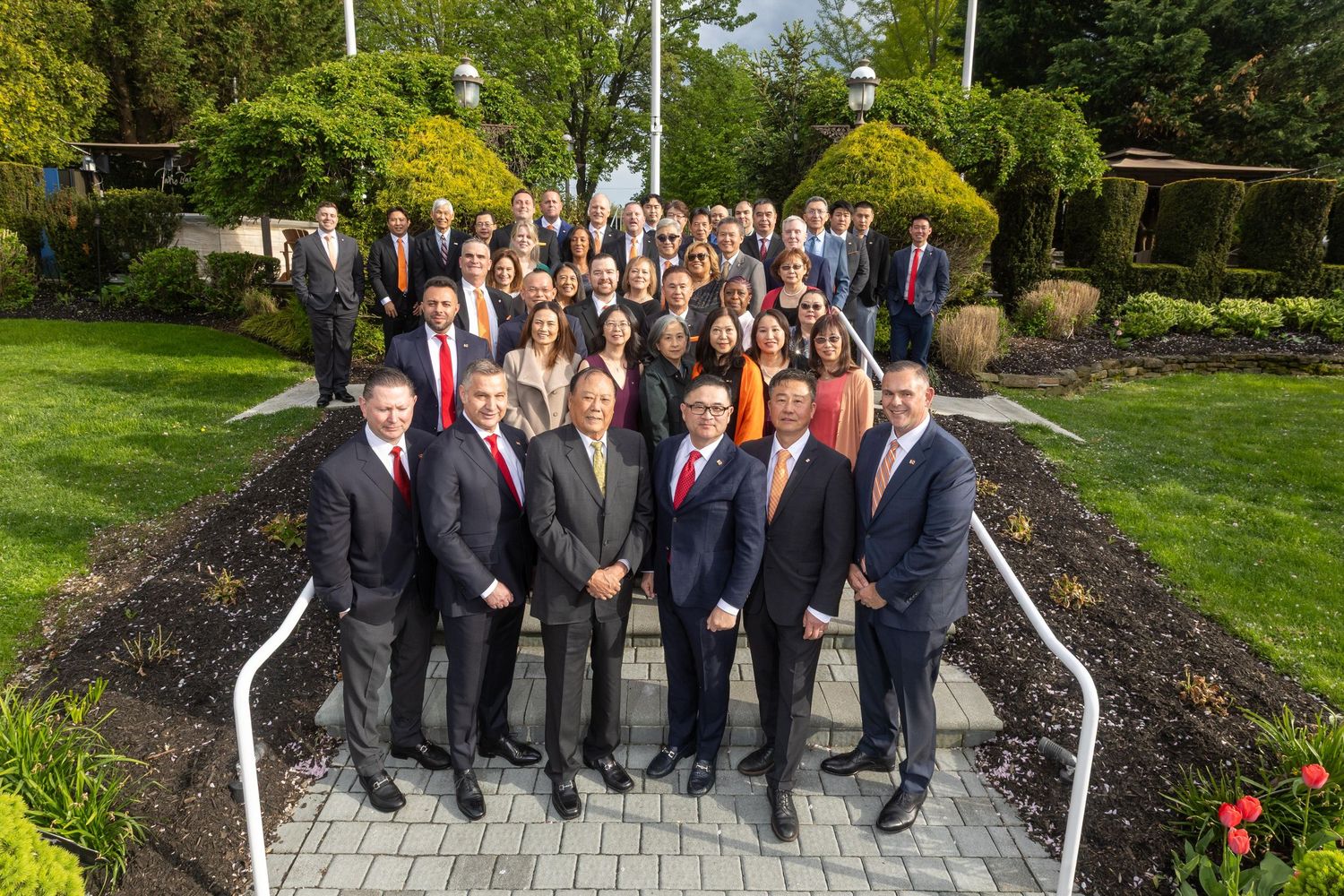 Large professional group photo of business team in formal attire posing outdoors on steps surrounded by greenery.