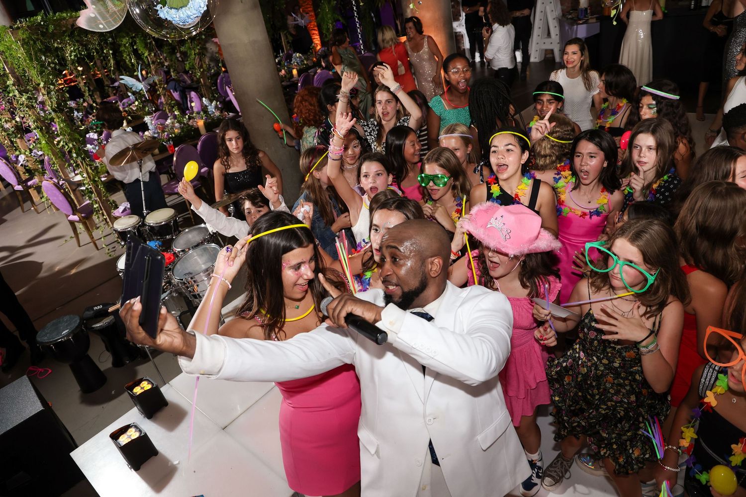 Energetic party crowd celebrating with colorful confetti, a man in white suit performing at a festive indoor event.