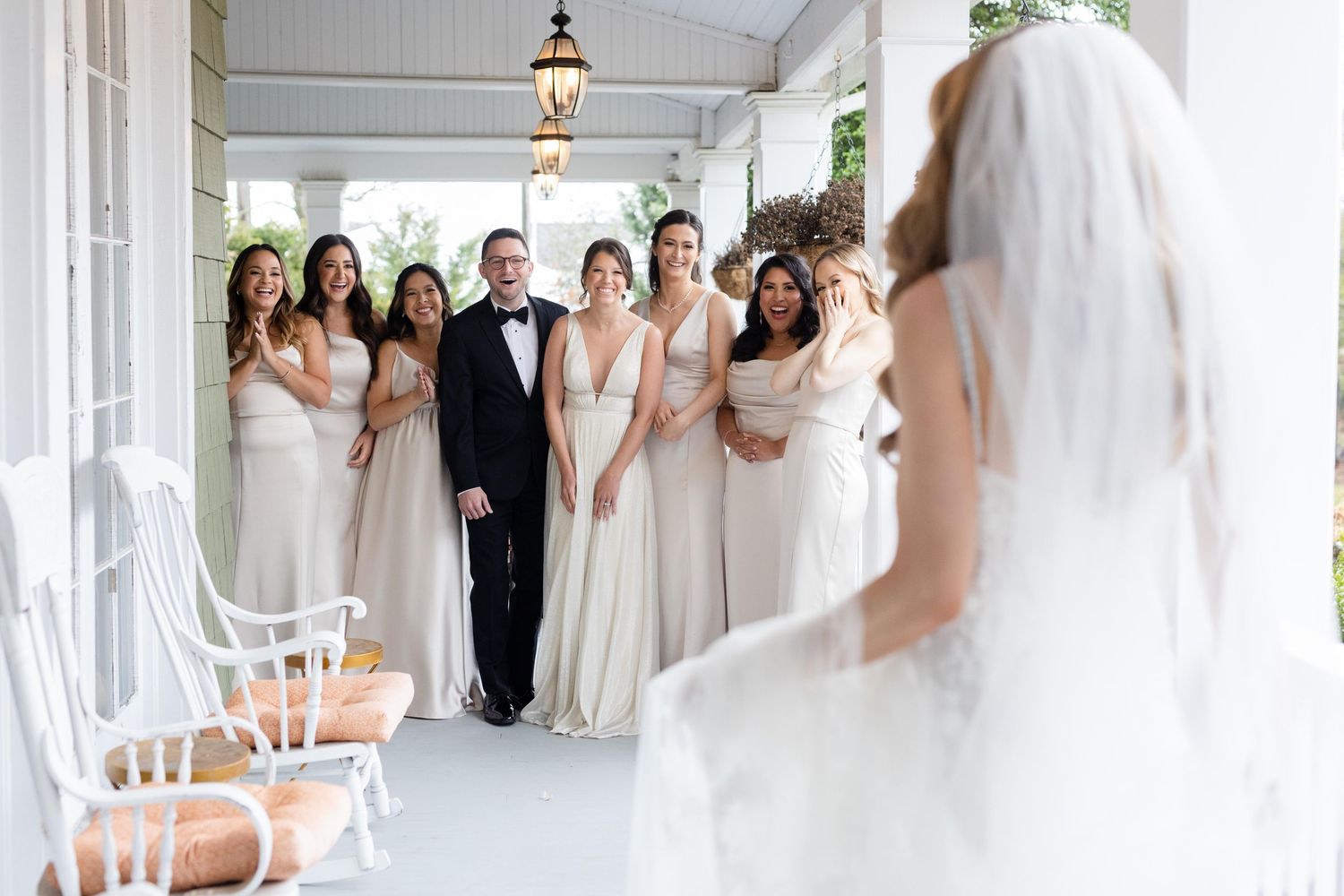 Wedding party in light colored dresses watches the bride's first reveal on a white porch.
