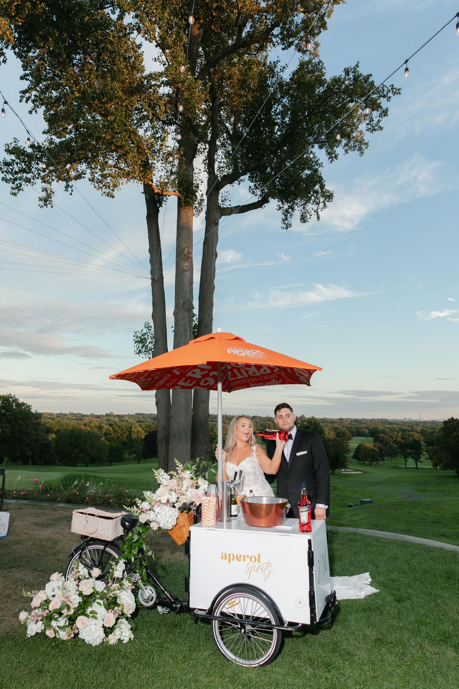 Wedding ice cream cart with orange umbrella decorated with flowers on scenic golf course.