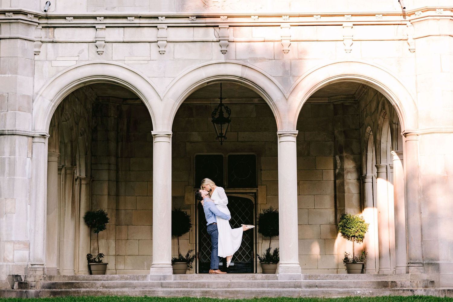 A romantic embrace under elegant stone archways with potted plants and hanging lanterns at sunset.