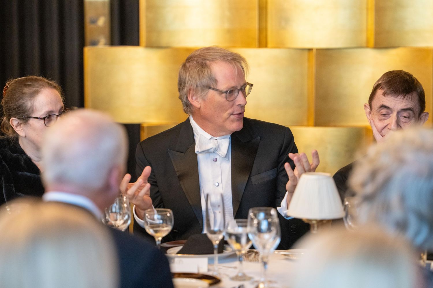 Elegant man in tuxedo gestures while speaking at a formal fundraising gala dinner.