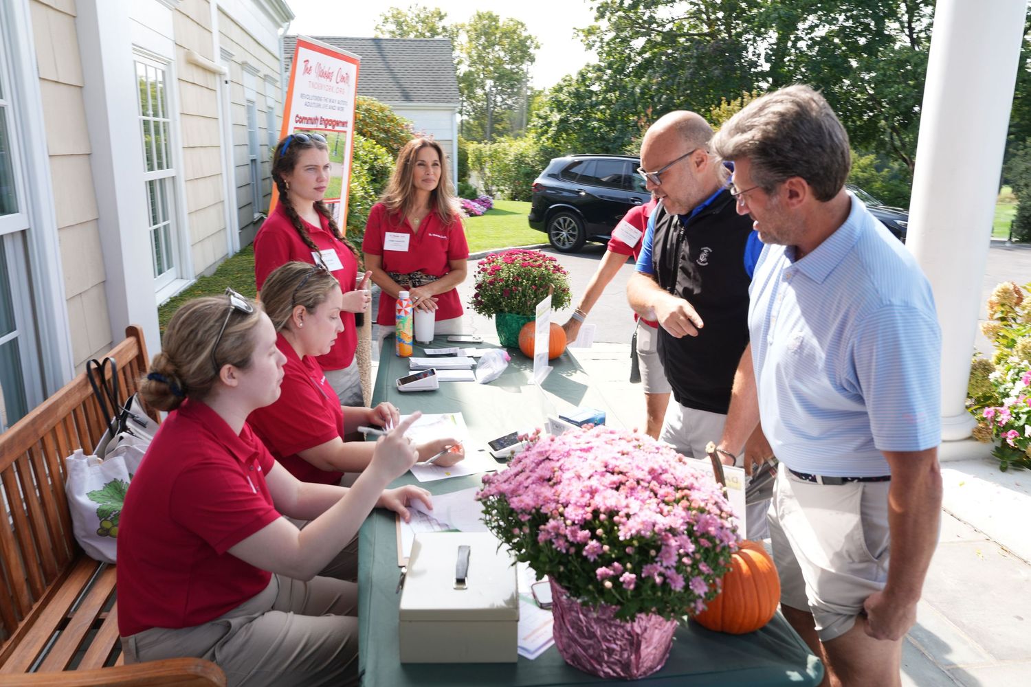 Volunteers in red shirts manage a registration table outdoors at a charity event with pink flowers and a pumpkin.