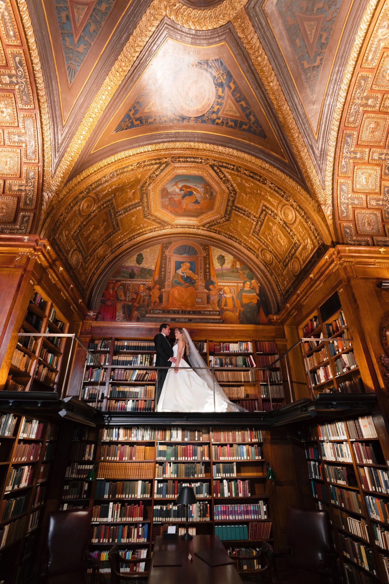 Wedding couple dances beneath ornate library ceiling with gilded details and bookshelves.