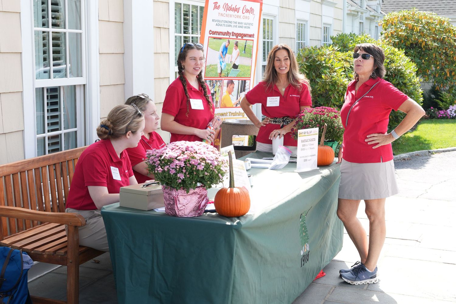 Four women in red polo shirts staff a registration table with flowers and pumpkins at a golf event.