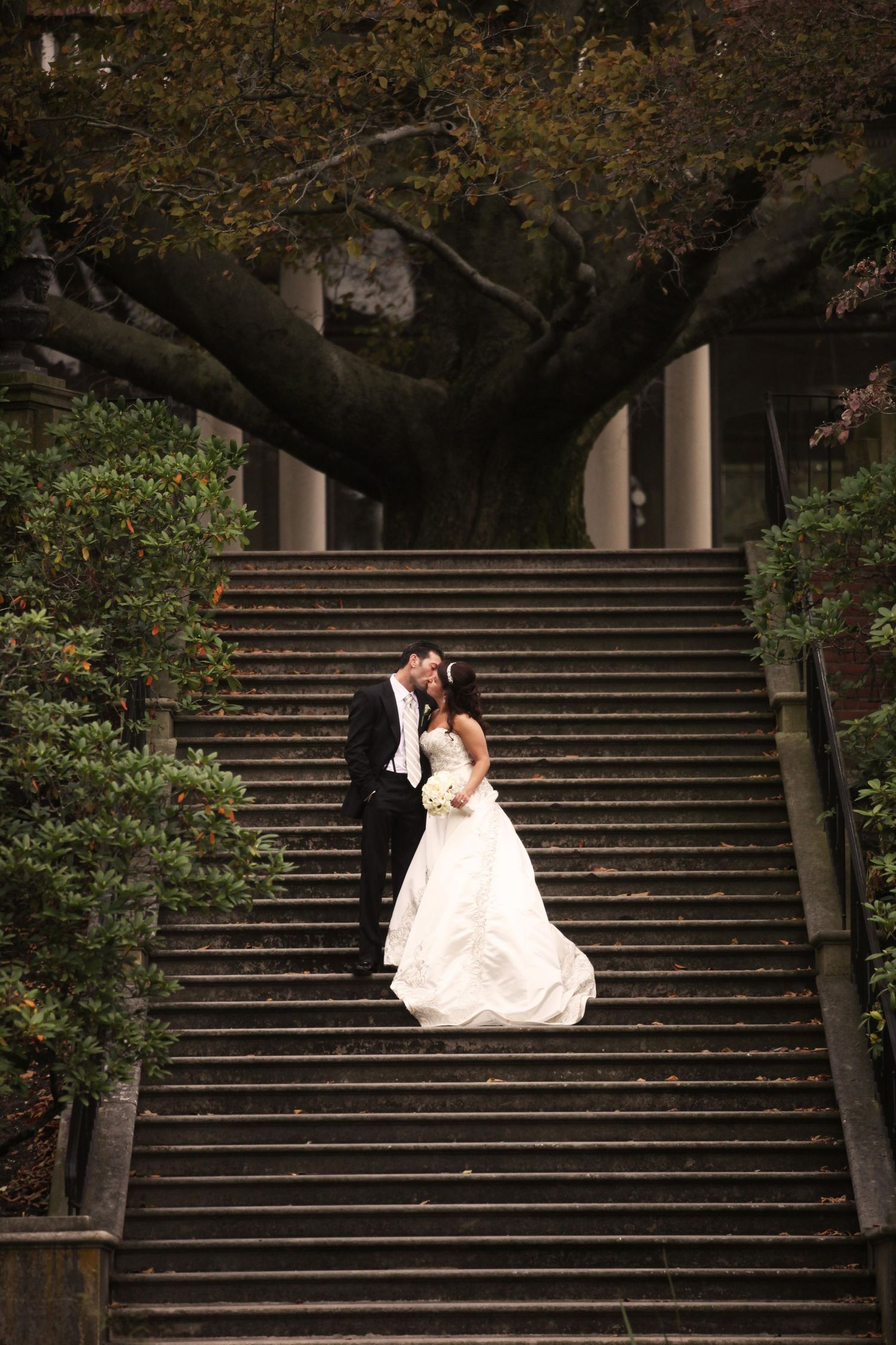 Wedding couple poses dramatically on grand stone steps surrounded by trees at dusk.
