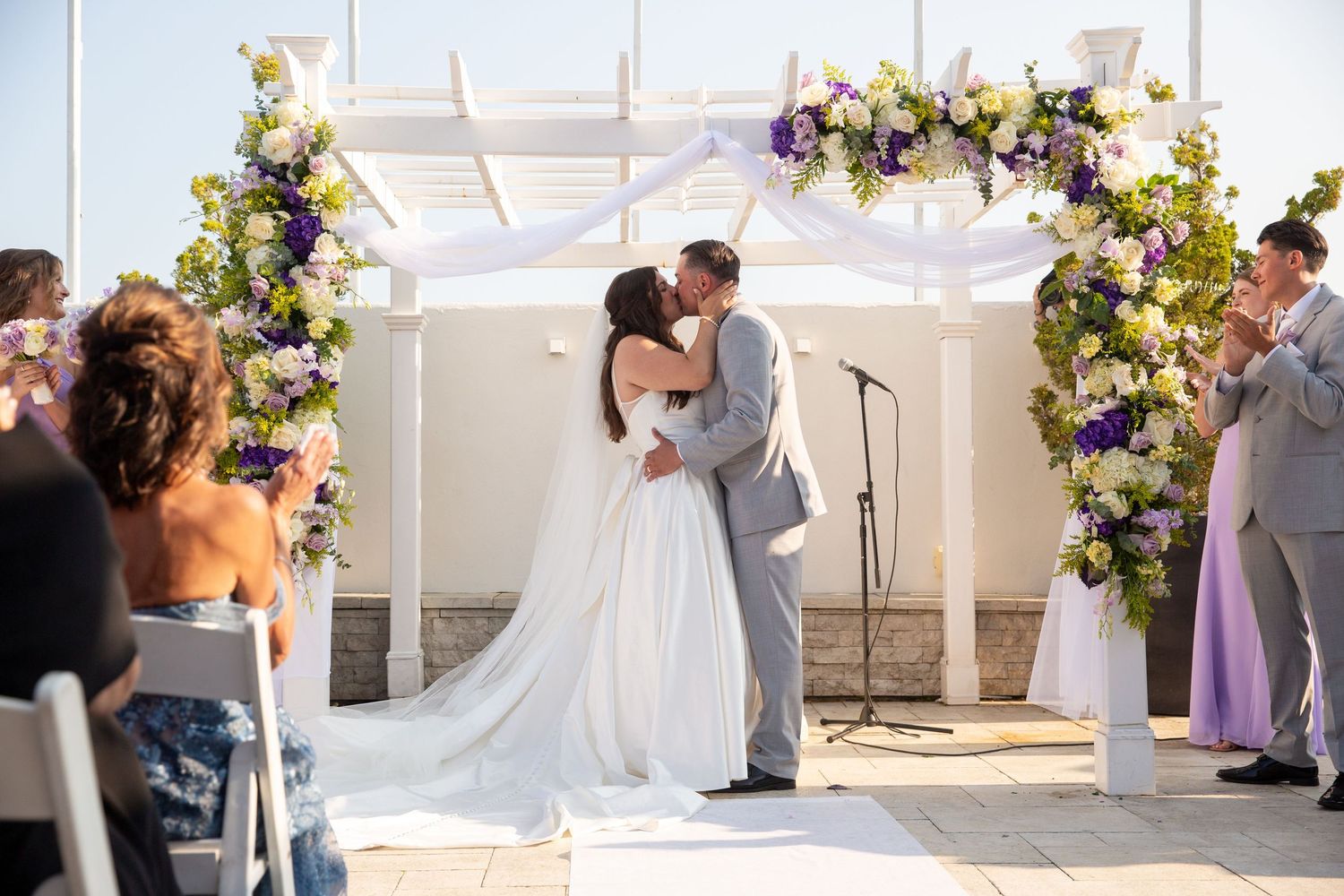 Wedding ceremony setup with purple and white floral arrangements against white backdrop.