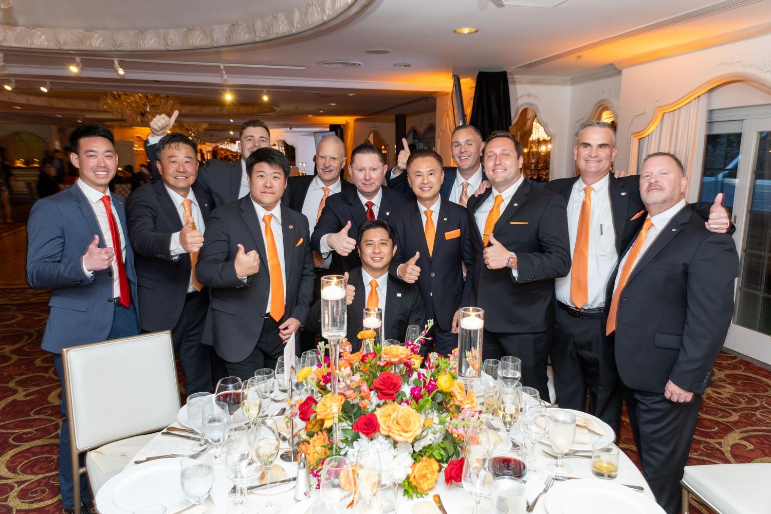 Group of men in suits with orange ties giving thumbs up at a formal dinner event with floral centerpiece.
